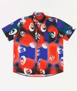 Camisa de manga corta con estampado multicolor de caras sonrientes sobre fondo rojo, azul y negro. Tiene cuello abotonado, bolsillo en el pecho y cierre frontal con botones.