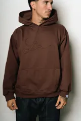 Hoodie oversize marrón oscuro con capucha y bolsillo canguro frontal. Presenta un logo bordado en tono gris claro en el pecho.