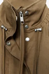 Chaqueta tipo bomber fluida color marrón, con cuello alto ajustable con cordones y stoppers, manga larga con elástico en los puños, bolsillos delanteros y bajo con pliegues. Cierre frontal con cremallera y botones a presión ocultos por solapa.