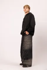 Pantalón de jean negro con proceso de lavado localizado y corte balloon fit. Presenta un diseño de recortes asimétricos, detalles de tachas metálicas y terminaciones desflecadas en el frente. Cuenta con bolsillos traseros clásicos y un calce holgado.