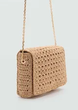 Bolso pequeño tipo bandolera confeccionado en tejido de fibra natural con diseño de espiga. Presenta detalles de tachuelas metálicas en los bordes, cierre de solapa con botón magnético y asa larga de cadena metálica dorada.