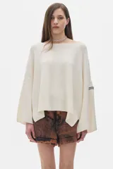 Buzo tejido de punto en color crudo, con cuello redondo, corte holgado y mangas largas amplias. Presenta un detalle de etiqueta en la manga.