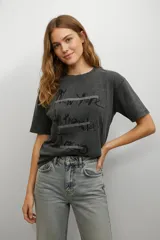 Remera de algodón con efecto lavado, cuello redondo y mangas cortas. Presenta un estampado gráfico con nombres de ciudades y detalles de apliques brillantes horizontales.