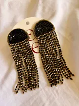 Aros colgantes de diseño semicircular bordados con mostacillas negras, de los cuales se desprenden múltiples hileras de cuentas negras y doradas.