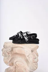 Sandalia negra de plataforma gruesa con dos tiras anchas ajustables con hebilla. Una de las tiras tiene un estampado de vaca blanco y negro.