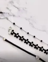 Set de tres gargantillas tipo choker en color negro. Incluye una cadena fina con cuentas negras, una gargantilla de flores negras con centro brillante y una cinta negra lisa con un dije circular central.