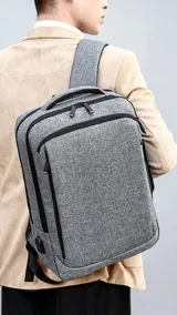 Mochila gris con detalles en negro, con doble bolsillo frontal con cierre, compartimento acolchado para laptop, puerto USB externo, asa de mano y faja trasera para acoplar a la manija de la valija.