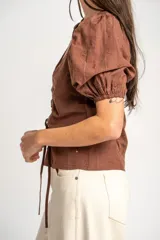 Blusa marrón de algodón con bordados sutiles, escote en V, mangas cortas abullonadas y detalle de fruncido con cordón ajustable en el centro del frente.