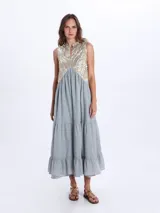 Vestido largo sin mangas, de color negro con la parte superior bordada en tonos dorados y negros con patrones geométricos. Presenta escote en V y falda con volados escalonados.