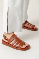 Sandalia estilo cangrejera confeccionada en cuero vacuno color suela, con suela plana de 3 cm y cierre mediante hebilla lateral.