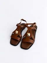 Sandalias planas de cuero color marrón, con punta cuadrada, tiras con recortes geométricos y aplique metálico dorado en la parte frontal. Cuenta con tira ajustable al tobillo con hebilla dorada.