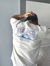 Remera de manga corta color beige jaspeado, con un estampado gráfico en la espalda que incluye el texto "TAS LOCO" y "SUNNY WITH A CHANCE OF STOKE" con un diseño de una persona en el agua con una ballena.
