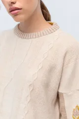 Sweater de punto color beige con cuello redondo acanalado, diseño de trenzas en el frente y bordados artesanales en las mangas.
