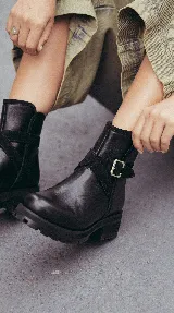 Bota corta de cuero negro con suela track de goma, detalle de tira cruzada con hebilla metálica lateral y paneles de gamuza en contraste.
