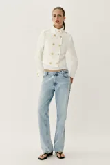 Chaqueta corta blanca de estilo militar, con cuello subido, manga larga y trabillas desflecadas en los hombros. Cuenta con cierre frontal cruzado decorado con botones metálicos dorados.
