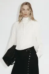 Camisa de manga larga color blanco hueso, con cuello camisero clásico y cierre frontal de botones. Presenta un corte recto y un bajo redondeado.