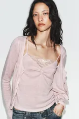 Chaqueta de punto color beige con escote en pico y manga larga, que incluye un top interior integrado con detalle de encaje a tono. Cierre frontal mediante botones.