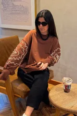 Buzo de manga larga color marrón, con cuello redondo y puños largos. Presenta un bordado lineal en color crema que simula formas orgánicas o florales en las mangas.