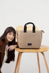 Cartera tipo tote de diseño capitoneado en color marrón claro, con doble asa de mano negra y bolsillos laterales. Presenta un diseño acolchado con líneas verticales en el frente y rombos en los laterales, e incluye una etiqueta decorativa con logo.