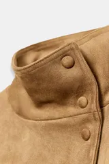 Chaqueta de gamuza sintética color marrón, con cuello subido, cierre frontal cruzado con botones y mangas largas anchas. Cuenta con bolsillos delanteros y cinturón ajustable del mismo tejido.