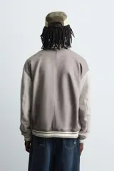 Cazadora estilo varsity o bomber de hombre, color gris oscuro con efecto ante, mangas en tono gris claro y puños, cuello y bajo con rib elástico a rayas blancas y grises. Presenta un parche con la letra 'R' en el pecho derecho y un bordado con texto en el pecho izquierdo.