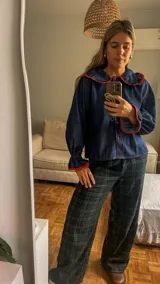 Camisa de jean azul oscuro con cuello estilo marinero y puños con volados, ambos con ribetes en color rojo. Presenta cierre frontal con botones rojos.