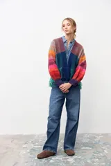 Chaqueta de tejido de punto con diseño multicolor a rayas diagonales en tonos cálidos y fríos. Presenta un panel frontal central en color azul sólido con flecos en el ruedo y puños tejidos en color violeta.