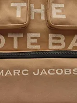 Bolso tipo tote Marc Jacobs modelo The Cargo Canvas Large Tote Bag, color tostado, confeccionado en lona con múltiples bolsillos delanteros con solapa y cierre, detalle de anilla metálica y estampado gráfico con el nombre de la marca. Cuenta con asas dobles para el hombro.