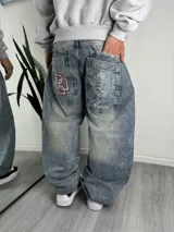 Jeans baggy de denim con efecto acid wash y lavado desgastado, de corte holgado. Presenta amplios bolsillos traseros con bordados contrastantes, incluyendo un diseño geométrico en rojo y blanco en un bolsillo y un motivo de querubín con la inscripción "Ameri" en el otro.