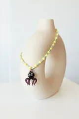 Collar corto con cuentas esféricas amarillas y cuentas cilíndricas a rayas blancas y verdes, con un dije de vidrio en forma de pulpo negro y morado.