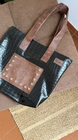 Bolso tote negro con textura símil cuero de cocodrilo y bolsillo frontal marrón con tachas plateadas. Tiene doble asa de hombro marrón y cierre superior.
