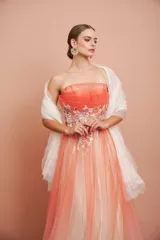 Vestido largo de fiesta, strapless, con busto drapeado y falda de tul con degradé de color coral a blanco. Presenta bordados florales en el corpiño.