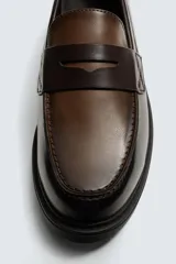 Mocasín de cuero marrón oscuro con antifaz en tono camel. Presenta horma redonda y suela track gruesa de goma negra.