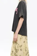 Camiseta de algodón color blanco hueso, de corte boxy y holgado, con escote redondo y mangas cortas. Presenta un pequeño logo en relieve de color rojo en el área del pecho.