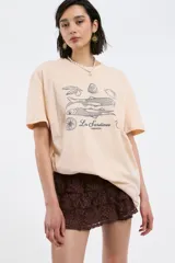 Remera de algodón color crema con corte oversize, cuello redondo y mangas cortas. Presenta un estampado frontal en color azul oscuro con motivos de sardinas, limones y aceitunas, junto con la inscripción 'Les Sardines Pimentón'.