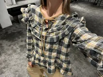 Camisa de manga larga con estampado a cuadros en tonos gris, negro y mostaza. Presenta volados en la parte superior delantera y en los hombros, con un diseño de corte holgado y lazos decorativos en el cuello.