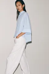 Camisa Zara Woman Collection de algodón con estampado de rayas verticales blancas y celestes. Presenta cuello perkins, mangas tres cuartos y un diseño asimétrico con cierre frontal cruzado mediante botones.