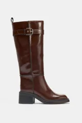 Bota alta de cuero sintético color marrón oscuro, con caña recta y punta cuadrada. Presenta pespuntes a contraste en las uniones y un detalle de correa con hebilla metálica en la parte superior. Posee un tacón de bloque de altura media.
