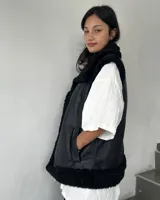 Chaleco estilo gamulán de cuero sintético marrón con interior y bordes de corderito sintético color crema. Presenta cierre frontal con botones a presión y bolsillos laterales.