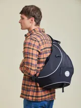 Mochila tipo bandolera (sling bag) de color negro con detalles de ribetes blancos. Presenta un diseño ergonómico de una sola correa, bolsillo frontal con cierre y logo circular de la marca Rusty. Incluye hebilla de ajuste lateral.