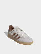 Zapatillas Adidas Handball Spezial de gamuza en tono gris claro, con las icónicas tres tiras laterales y refuerzo en el talón con textura de piel de serpiente en color marrón. Presenta suela de goma color caramelo y detalles dorados en el logo lateral y la lengüeta.