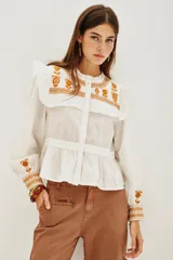 Camisa de algodón celeste con detalles de volados en los hombros y bordados en color negro en el pecho y las mangas. Presenta cierre frontal con botones y puños ajustados.