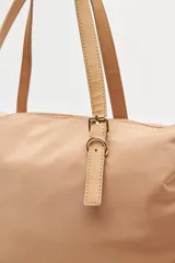Cartera tipo tote de nylon negro, con diseño amplio y liviano. Presenta asas superiores fijas con detalles de tiras regulables y herrajes metálicos dorados. Incluye cierre principal y un pequeño logo metálico en la parte inferior frontal.