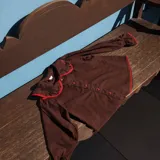 Camisa de color marrón chocolate con cuello Peter Pan y puños con volados, ambos detalles resaltados con un ribete en color rojo. Presenta cierre frontal con botones rojos.