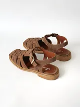 Sandalias estilo cangrejera color marrón, confeccionadas en cuero con diseño trenzado en la parte delantera, tira ajustable con hebilla metálica en el tobillo y suela plana.