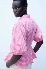 Camisa de popelín a rayas rosadas y blancas, con cuello solapa, mangas abullonadas por debajo del codo con terminación elástica, cintura entallada y bajo en evasé. Cierre frontal con botones ocultos.
