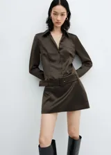 Vestido corto de satén color marrón oscuro, con cuello camisero, cierre frontal de botones y mangas largas con puños abotonados. Incluye un cinturón desmontable con hebilla a tono y cierre lateral con cremallera invisible.
