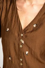 Blusa de viscosa color marrón, con escote en V, mangas cortas abullonadas y cierre frontal con botones. Cuenta con pequeños detalles bordados distribuidos por la prenda.