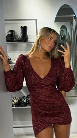 Vestido corto de fiesta confeccionado en tejido de lurex color bordó con destellos brillantes. Presenta un escote en V profundo, mangas largas abullonadas y un diseño ajustado al cuerpo con un corte cruzado en el ruedo.