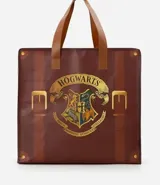 Bolsa tipo shopper de material plástico resistente, con diseño estampado que simula una maleta clásica con correas y hebillas. Presenta el escudo de Hogwarts en el centro y cuenta con asas dobles para llevar al hombro.